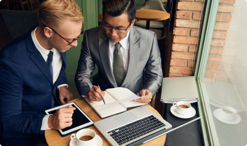 Overhead view of two professional business consultants in formal suits reviewing documents, a laptop, and a tablet while discussing project strategy at a small cafe table.
