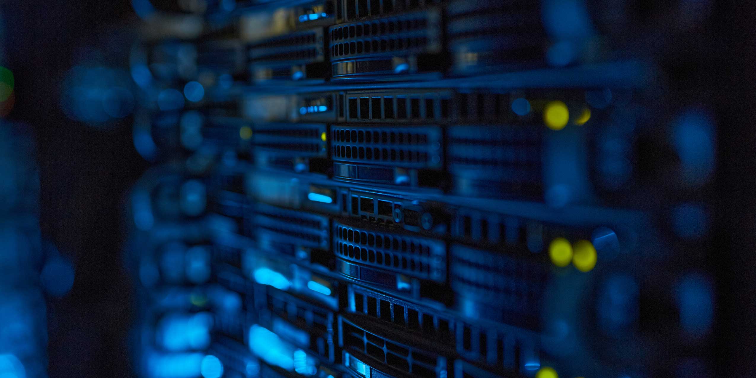 A selective-focus shot of stacked network server blades in a dark data center, illuminated by blinking blue status lights and faint yellow indicator bulbs.