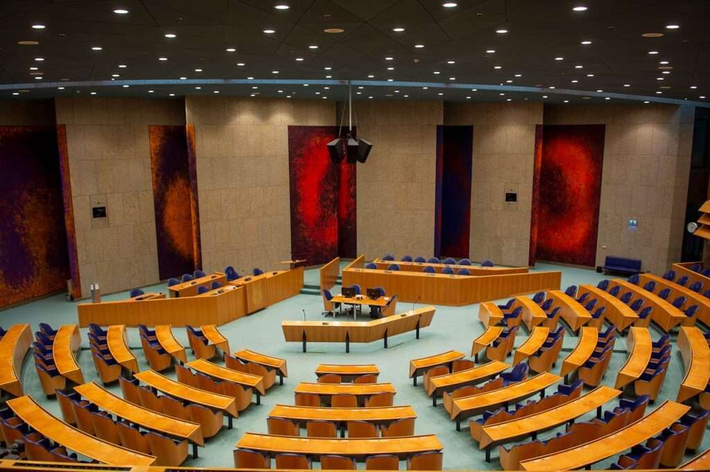 A wide view of a large, circular legislative auditorium or conference center featuring curved rows of polished wooden desks, blue cushioned chairs, and abstract red wall paneling beneath a starry grid of ceiling lights.