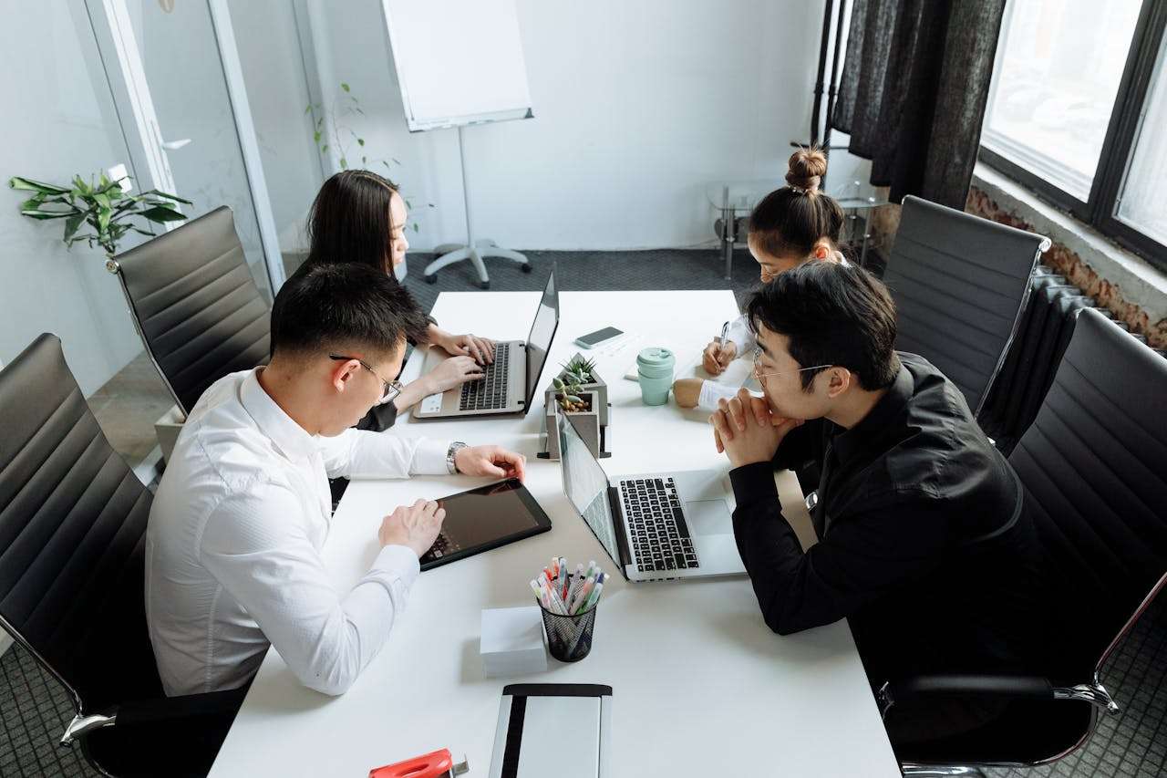 A high-angle shot of four corporate professionals collaborating around a white conference table, using multiple laptops and a tablet to review project strategies.