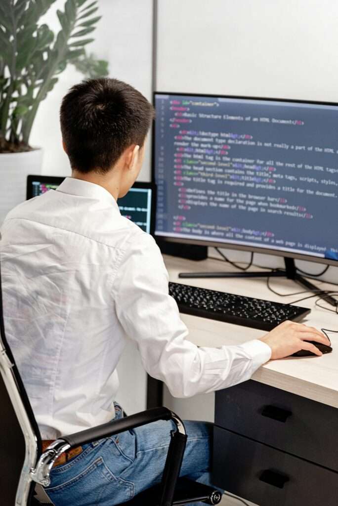 A web developer in a white shirt sits at a desk looking at a monitor displaying clean HTML and CSS code detailing the basic structural elements of a web document.