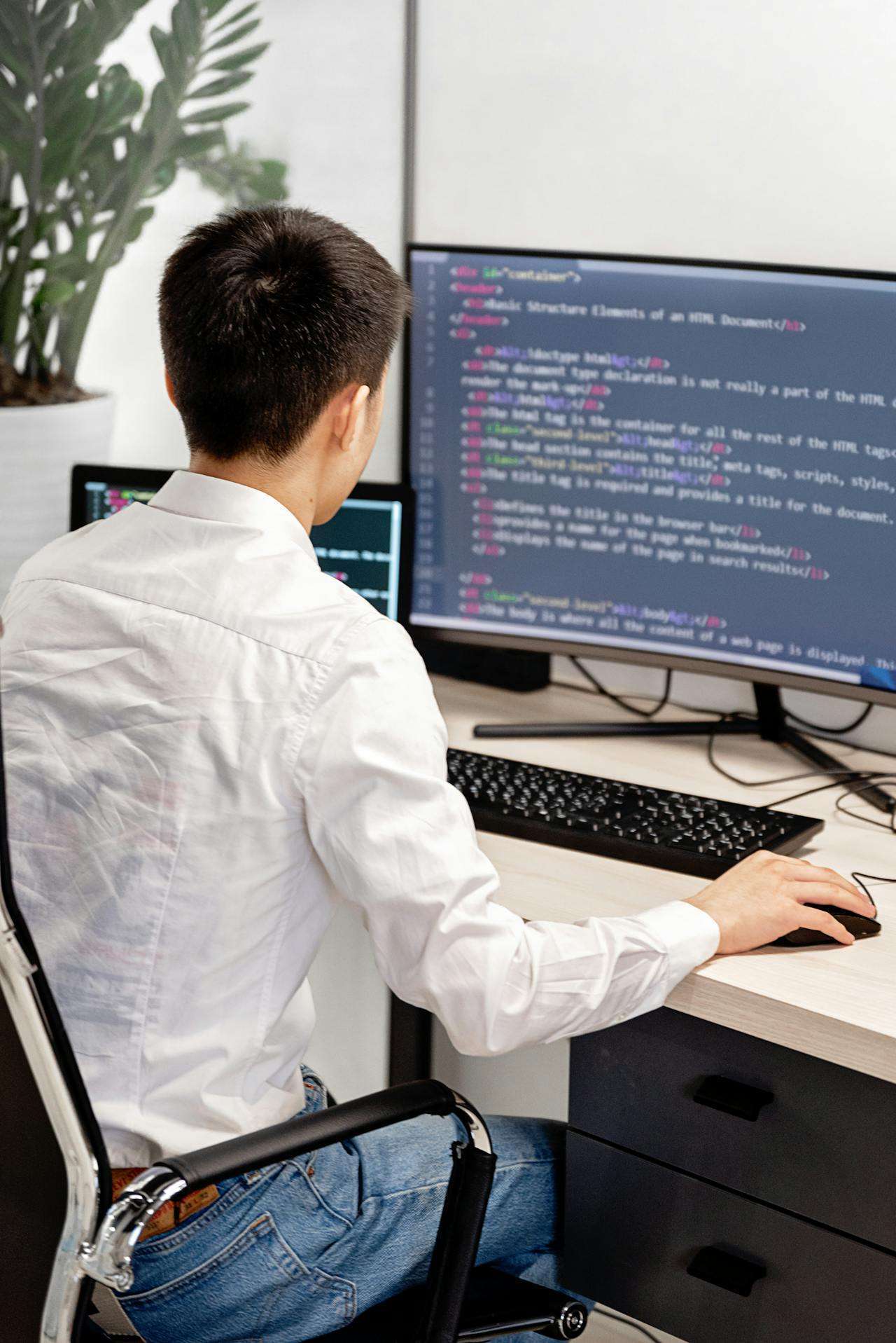 A web developer in a white shirt sits at a desk looking at a monitor displaying clean HTML and CSS code detailing the basic structural elements of a web document.