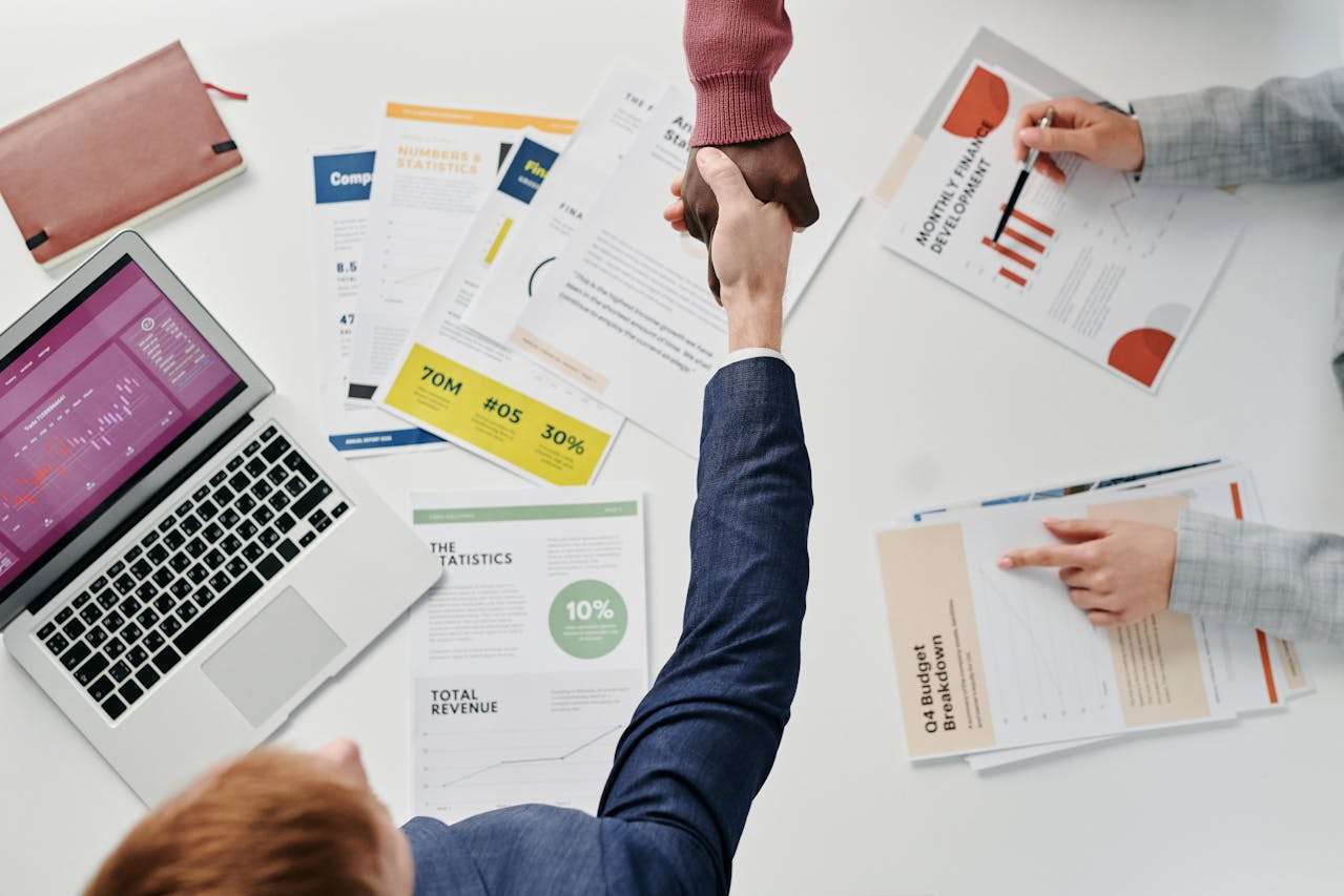 An overhead shot of two professionals shaking hands over a white office table filled with data analysis charts, a laptop showing statistics, and financial performance reports.
