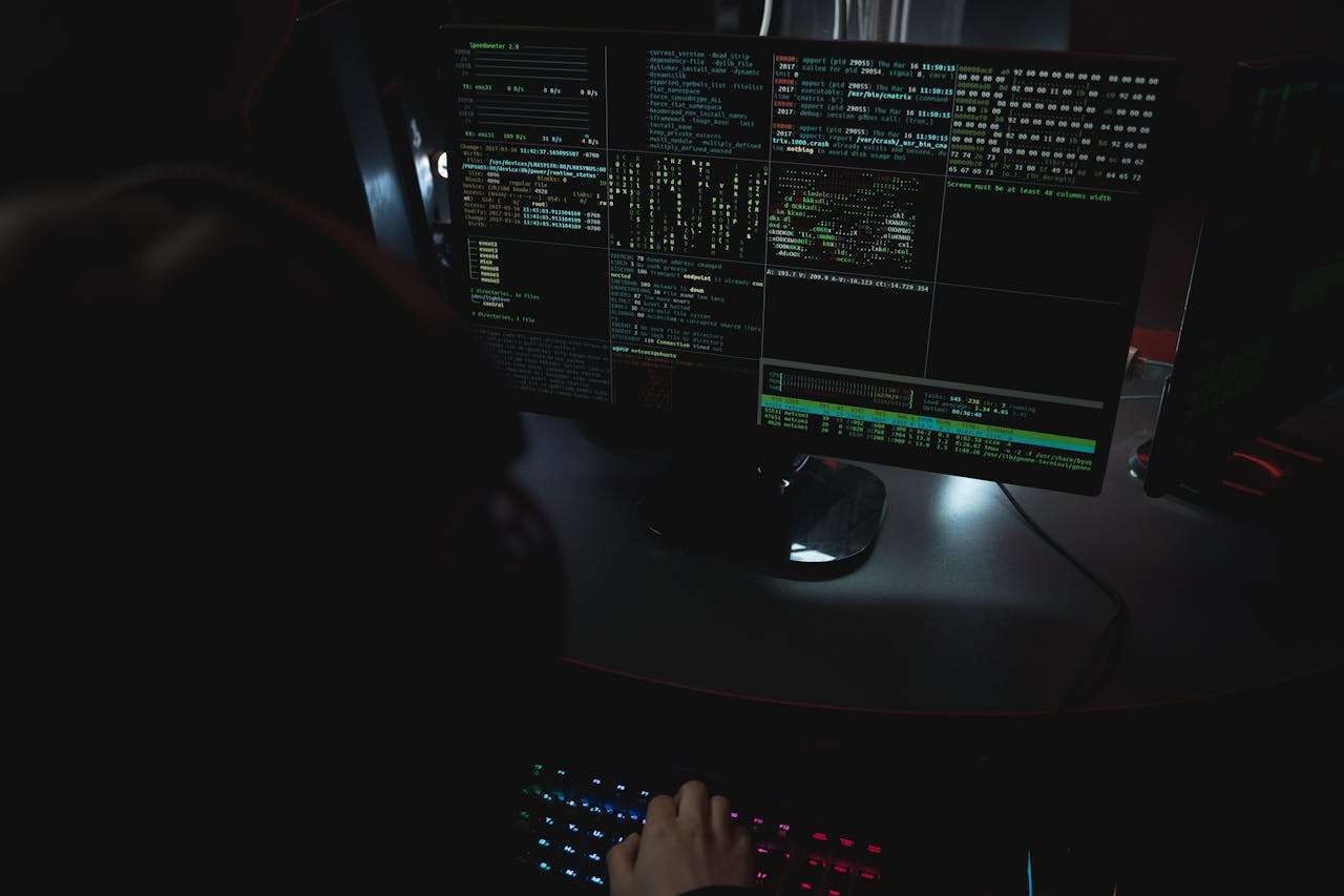 A dark room perspective of dual monitors displaying detailed server log data, code strings, and command-line terminal readouts during a technical system audit.